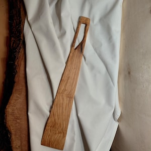 Może przedstawiać: Drewniana szpatułka z długim, smukłym uchwytem i prostokątnym ostrzem. Uchwyt ma unikalny, wycięty wzór. Szpatułka wykonana jest z jasnobrązowego drewna z widocznym wzorem słojów, co sugeruje, że jest wykonana z naturalnego materiału.