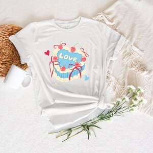 May include: White t-shirt featuring a heart-shaped cake design with the word "LOVE" in blue, adorned with cherries and red ribbons. The cake is surrounded by yellow frosting and small hearts. A casual, everyday tee.