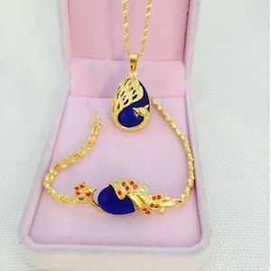 May include: A gold-colored jewelry set featuring a pendant necklace and bracelet. The pendant and bracelet each have a large, teardrop-shaped, deep blue stone. The gold setting has a decorative openwork design and is accented with red details. The set is displayed in a pink box.