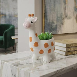 May include: A white ceramic giraffe planter with orange spots, holding a green succulent. The giraffe has a pink nose and ears. The planter is on a marble surface.