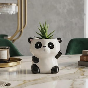 May include: A white ceramic panda planter with black accents, holding a green succulent plant. The panda has black ears, eyes, and paws. The planter is sitting on a marble table.