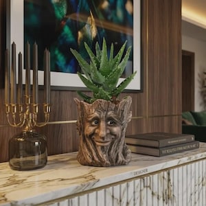 May include: A whimsical, brown tree-stump planter with a smiling face, holding a green aloe vera plant. A gold candelabra with dark candles sits to the left. The planter is on a marble surface.