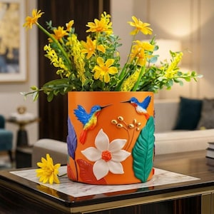 May include: An orange ceramic flower pot with a hummingbird and floral design. The pot features a white flower, colorful leaves, and gold accents. Yellow flowers and greenery are arranged inside the pot, creating a vibrant display.