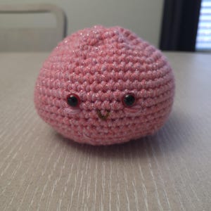 May include: A round, pink crocheted plush toy with a subtle shimmering thread woven throughout. The object features two small, dark, glossy safety eyes and a tiny, embroidered brown V-shaped mouth, with faint pink stitched details below each eye.