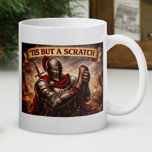 May include: White ceramic mug with a medieval knight design. The knight is in full armour, with a sword and the text "Tis But a Scratch" above. The mug has a white handle and a glossy finish. The image is set against a festive background.