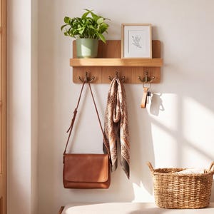 May include: A light brown wooden wall-mounted shelf featuring three bronze-toned double hooks. The shelf holds a green potted plant and a framed line drawing. A brown leather crossbody bag, a patterned scarf, and a keychain hang from the hooks.