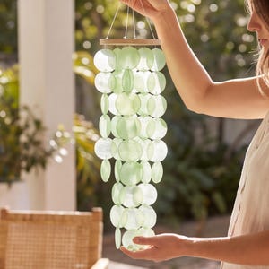 Peut inclure: Un mobile suspendu composé de multiples disques circulaires translucides vert pâle, fixés à un cadre en bois circulaire par de fines ficelles blanches. Les disques sont disposés en brins verticaux, créant une structure en cascade superposée.