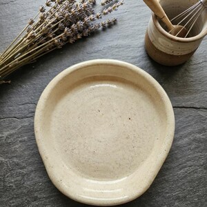 May include: A round, cream-colored ceramic spoon rest with a speckled finish and a slightly raised rim. The spoon rest has a smooth, glossy glaze and a simple, functional design intended for holding kitchen utensils while cooking.
