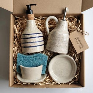 Puede incluir: Un juego de cocina en una caja de cartón que contiene un dispensador de jabón de cerámica moteada con rayas azules, una aceitera de cerámica moteada con boquilla metálica, un soporte para esponja con esponja azul y un plato pequeño.