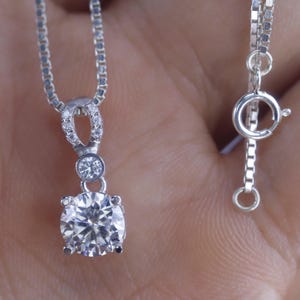 May include: A silver pendant necklace featuring a large, round, clear gemstone as the focal point. The pendant has a smaller gemstone above the main stone and is accented with smaller stones. The necklace chain is also silver.