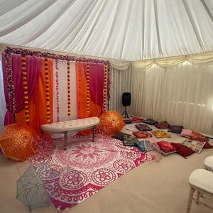 Può includere: Uno spazio per eventi decorato con un soffitto a tenda bianco, uno sfondo rosa e arancione adornato con fiori, una panca, un tappeto fantasia e cuscini colorati. Anche ombrelli decorativi sono disposti sul pavimento.