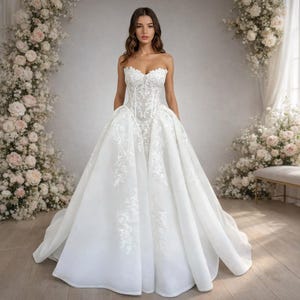 Puede incluir: Un vestido de novia blanco sin tirantes con intrincados detalles de encaje en el corpiño y la falda. El vestido presenta una falda amplia y fluida y un escote corazón. El fondo incluye arreglos florales y un banco.