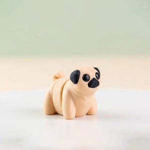 May include: A small, tan pug figurine with black accents on its ears, nose, and eyes. The pug is in a sitting position, with its body segmented. The background is a gradient of light green and beige.