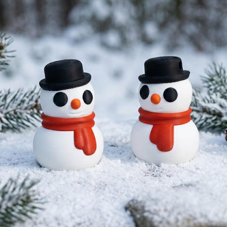 May include: Two small snowman figurines with black top hats, orange noses, and red scarves. The snowmen have black button eyes and are set in a snowy environment with evergreen branches in the background. The snowmen are a festive holiday decoration.