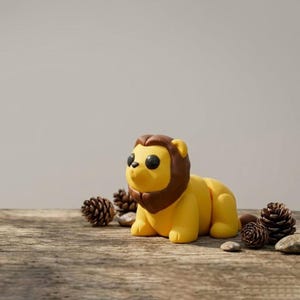 May include: A yellow lion figurine with a brown mane and black eyes, resting on a wooden surface. The figurine is surrounded by pine cones and small stones, creating a natural, earthy aesthetic. The lion is a cute, stylized representation.