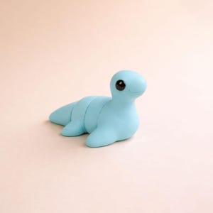 May include: A light blue, handmade clay sculpture of a sea creature with a smiling face and large black eyes. The aquatic animal has a segmented body and flipper-like appendages, resting on a light pink surface.