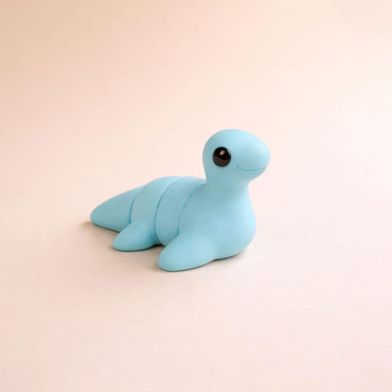 May include: A light blue, handmade clay sculpture of a sea creature with a smiling face and large black eyes. The aquatic animal has a segmented body and flipper-like appendages, resting on a light pink surface.