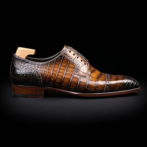 May include: A brown leather oxford shoe with a textured crocodile skin pattern, featuring a cap toe with decorative brogue detailing, dark brown laces, and a wooden shoe tree inserted into the heel area, set against a solid black background.