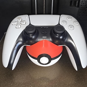 Puede incluir: Un mando blanco y negro de PlayStation 5 apoyado en un soporte rojo y blanco con el tema de la Poké Ball. El mando tiene joysticks negros y botones blancos. El soporte es rojo con un círculo blanco.