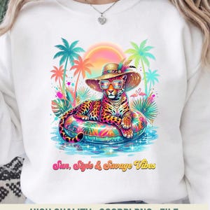 May include: White sweatshirt featuring a colorful illustration of a leopard wearing sunglasses and a sun hat, lounging on an inflatable ring in water. Palm trees and the text "Sun, Style & Savage Vibes" are also visible.