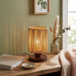 May include: A cylindrical table lamp featuring a shade made of vertical natural-colored woven rope, set upon a circular dark brown wooden base. The lamp emits a warm glow, illuminating the surrounding space with soft, vertical light patterns.