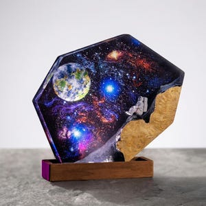 May include: A unique resin art piece depicting a vibrant galaxy scene with a seated astronaut. The artwork features a detailed Earth, stars, and nebulae, set against a wooden base. The piece is a striking decorative item.