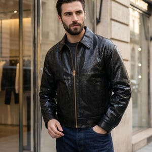 May include: A black jacket with a textured snakeskin-like pattern, featuring a classic collar, long sleeves, and a central gold-toned metal zipper closure. The garment is worn over a dark grey shirt and paired with dark blue denim jeans.