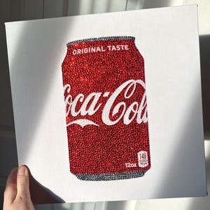 Puede incluir: Un diseño de lata de Coca-Cola roja, cubierto de brillantes adornos, sobre un lienzo blanco. Las palabras "Coca-Cola" están en escritura blanca, y los detalles de la lata incluyen "Original Taste" y "12oz".