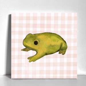 May include: A square art print featuring a watercolor illustration of a green frog with a large dark eye, positioned centrally against a soft light pink and white gingham checkered background pattern, creating a simple and clear visual composition.