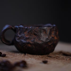 May include: A small, handcrafted ceramic coffee cup with a textured, dark brown exterior and a small handle. The cup has a rustic, organic appearance, with visible imperfections. Coffee beans are scattered near the cup.