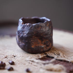 Może przedstawiać: Mały, ręcznie wykonany ceramiczny kubek o szorstkiej, teksturowanej powierzchni. Kubek jest ciemnobrązowy z nutą pomarańczu i stoi na teksturowanej, beżowej powierzchni. Kilka ziaren kawy jest rozrzuconych.