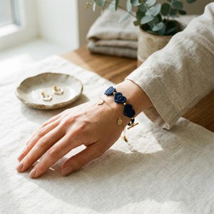 Op de afbeelding: Een gouden armband met kleine bedels en een marineblauwe, hartvormige gehaakte armband. De armband zit om de pols van een persoon. Op de achtergrond een klein keramisch schaaltje met gouden oorbellen.