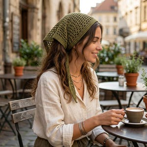 May include: An olive green crochet headscarf with a triangular shape and an openwork pattern. The headscarf is worn tied at the back of the head, with the pointed end draping down. It features a textured, knotted yarn construction in a solid color.