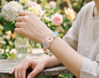 Puede incluir: Una pulsera de ganchillo con un diseño floral en tonos pastel, incluyendo rosa, morado, azul y verde. La pulsera se lleva en la muñeca, mostrando el patrón floral hecho a mano. El fondo es una escena de jardín borrosa.