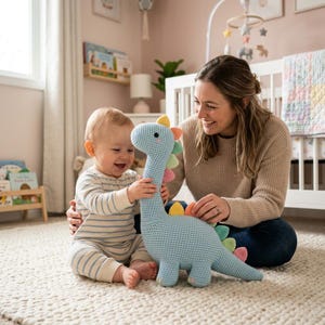 Könnte beinhalten: Ein hellblaues gehäkeltes Dinosaurierspielzeug mit bunten Stacheln. Der Dinosaurier hat einen langen Hals und ein lächelndes Gesicht. Das Spielzeug wird von einem Baby und einem Erwachsenen gehalten. Die Szene spielt in einem Kinderzimmer mit einem Kinderbett und einem Fenster.
