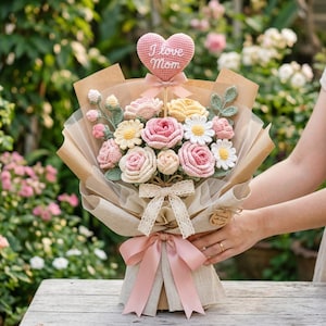 May include: A crochet bouquet with pink, yellow, and white flowers, wrapped in brown paper and tied with a pink ribbon. A pink heart-shaped sign with "I love Mom" is attached. The bouquet is a unique gift idea, perfect for Mother's Day.