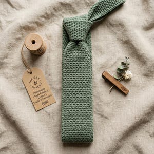 Può includere: Una cravatta all'uncinetto verde salvia fatta a mano. La cravatta è realizzata in cotone e ha un aspetto strutturato. Una bobina di filo di legno e un'etichetta con la scritta "THE OAK & THREAD Handmade | Sage Green | Cotton | Made in Vermont" sono accanto alla cravatta.