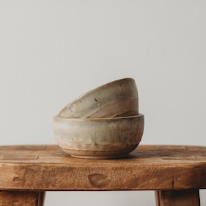 May include: Two handcrafted ceramic bowls, one stacked atop the other, with a rustic, earthy aesthetic. The bowls are a muted beige color with subtle variations. They rest on a weathered, wooden stool, showcasing a natural, minimalist design.