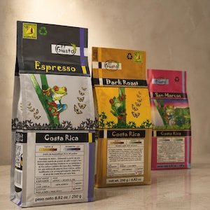Può includere: Tre sacchetti di caffè rettangolari etichettati El Gusto, con l'illustrazione di una rana dagli occhi rossi. I sacchetti sono neri, gialli e rosa, riportano Costa Rica, livelli di tostatura e un peso netto di 250 g. Quattro chicchi sono in primo piano.