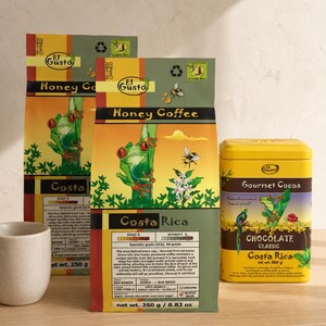 Può includere: Sono esposti tre pacchetti di "El Gusto Honey Coffee Costa Rica" e una scatola gialla di "Gourmet Cocoa Chocolate Classic Costa Rica". Le confezioni di caffè presentano un'illustrazione di una rana verde. La scatola di cacao ha un design di rana e uccello.