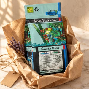 Op de afbeelding: Een lichtblauwe koffiezak met het label San Ramón Costa Rica en een illustratie van een roodoogmakikikker. De zak bevat tekst zoals 100% Arabica, 8,82 oz en 250 g. Het zit in bruin papier met een takje gedroogde lavendel en een leeg label.