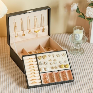 以下が含まれることがあります： 黒い持ち手と「But First, Coffee」のテキストが太字の黒で書かれた白いセラミックマグ。マグカップはぼやけた背景に置かれており、居心地の良い環境を示唆しています。