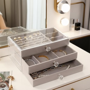 Puede incluir: Un organizador de joyas de acrílico transparente y terciopelo gris con tres cajones y un compartimento superior. Los cajones contienen varios collares, pulseras y anillos. El organizador tiene pomos de cristal y está sobre una superficie blanca.
