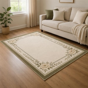 May include: Rectangular area rug with a floral design. The rug features a cream-colored center with a border of green and a floral pattern in the corners. The rug is placed on a hardwood floor in a living room setting.