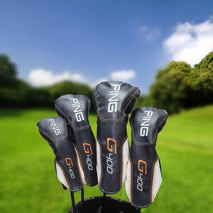 May include: A set of black and white golf club head covers with the brand name "PING" in white lettering. The covers have orange accents and the "G400" model name. The background shows a green golf course under a blue sky.