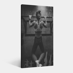 May include: Black and white photograph of a person doing a pull-up on a bar. The person is wearing a sports bra and leggings. The image is taken from behind, showcasing the person's back muscles. The background is a textured wall.