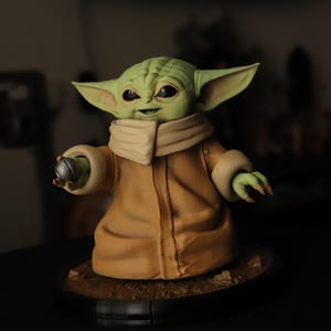 Puede incluir: Una figura de criatura de piel verde con grandes ojos oscuros y orejas puntiagudas, que viste una t&uacute;nica color canela con cuello alto y pu&ntilde;os. La figura sostiene un peque&ntilde;o objeto esf&eacute;rico plateado en una mano sobre una base circular.