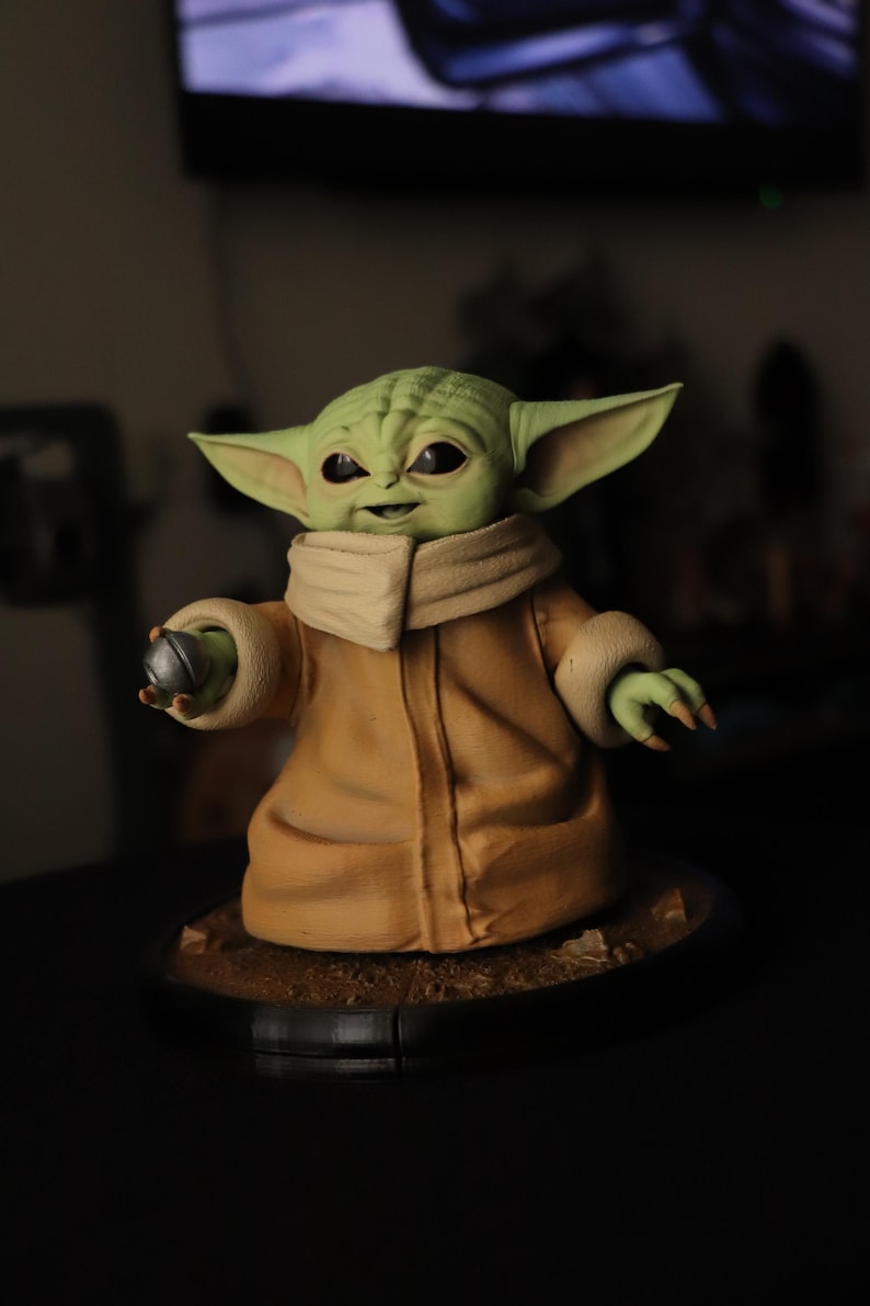 Puede incluir: Una figura de criatura de piel verde con grandes ojos oscuros y orejas puntiagudas, que viste una t&uacute;nica color canela con cuello alto y pu&ntilde;os. La figura sostiene un peque&ntilde;o objeto esf&eacute;rico plateado en una mano sobre una base circular.