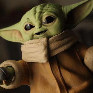 Puede incluir: Una figura de piel verde con grandes ojos oscuros, orejas puntiagudas y frente arrugada. El personaje viste una t&uacute;nica de color beige con un cuello grueso y texturizado alrededor del cuello y pu&ntilde;os a juego, mostrando un parecido detallado.