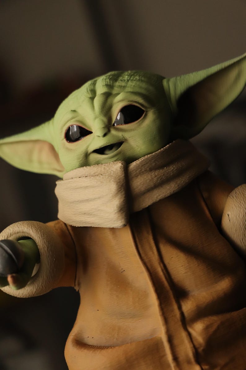 Puede incluir: Una figura de piel verde con grandes ojos oscuros, orejas puntiagudas y frente arrugada. El personaje viste una t&uacute;nica de color beige con un cuello grueso y texturizado alrededor del cuello y pu&ntilde;os a juego, mostrando un parecido detallado.